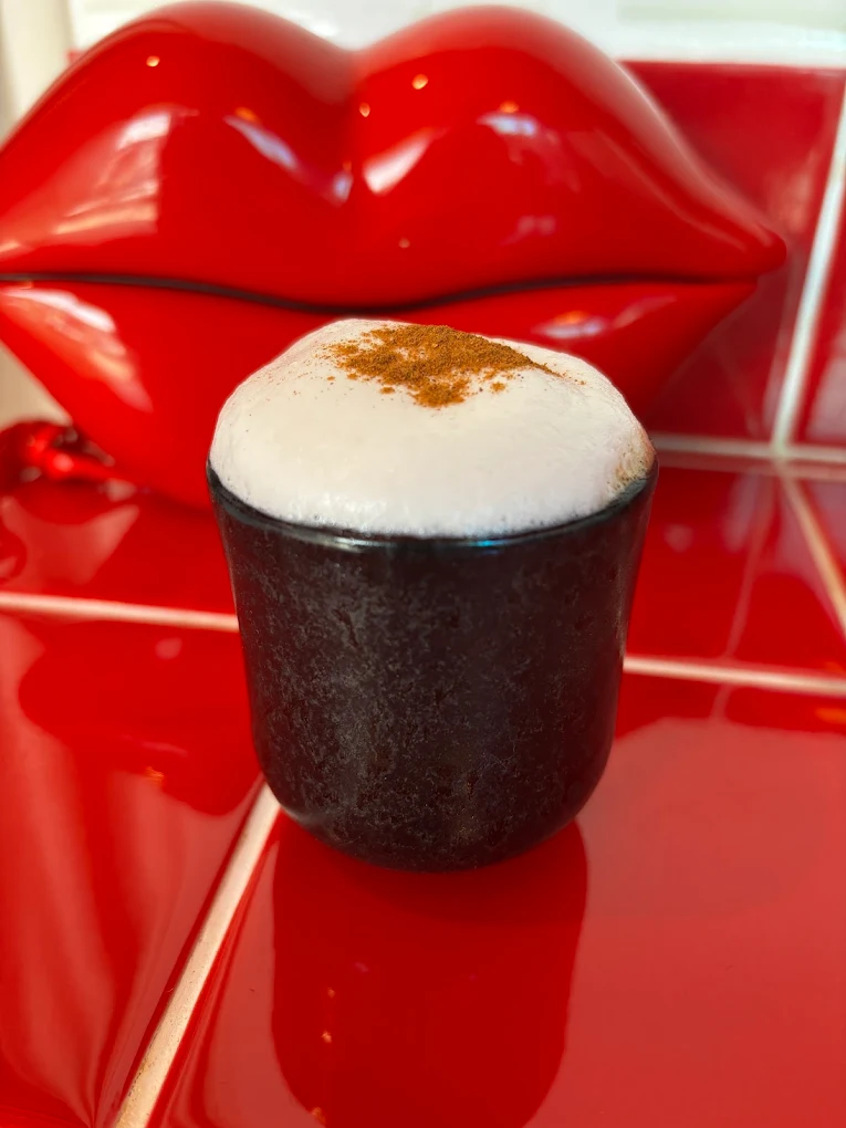 A foamy cappuccino in a black ceramic cup by a red lip sculpture